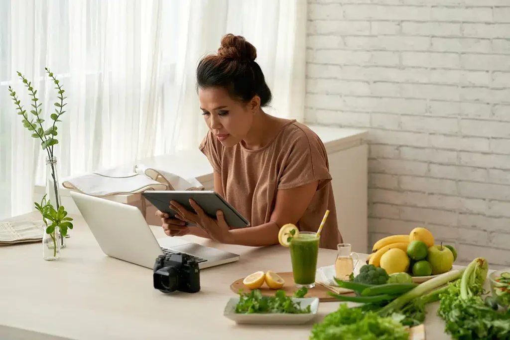 Woman in front of her laptop holding a tablet at a desk with fruits and vegetables, illustrating hybrid business models